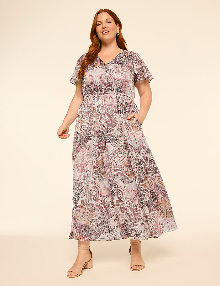 A woman with red hair wears a patterned maxi dress and beige heels against a plain, light-colored background.
