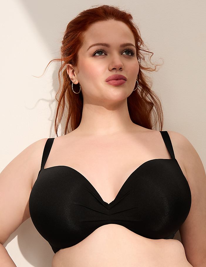 A woman with red hair wearing a black bra stands against a plain, light-colored wall.