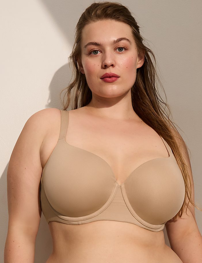 A woman wearing a beige bra stands against a plain, light-colored wall.