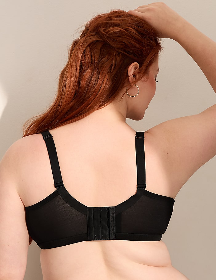 A woman with red hair wearing a black bra stands against a plain, light-colored background.