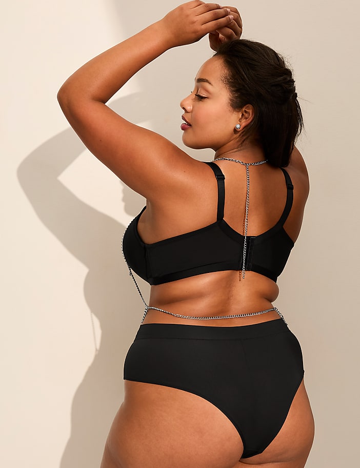 A woman in a black two-piece swimsuit with silver chain accents poses against a plain, light-colored wall.