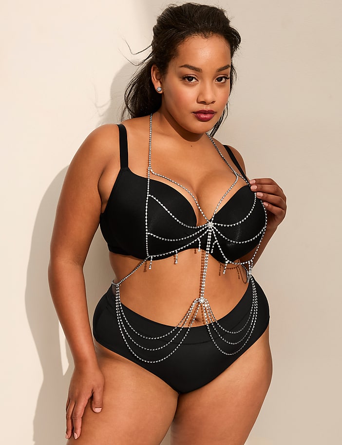 A woman wearing a black bikini adorned with silver beaded chains poses against a plain white wall.