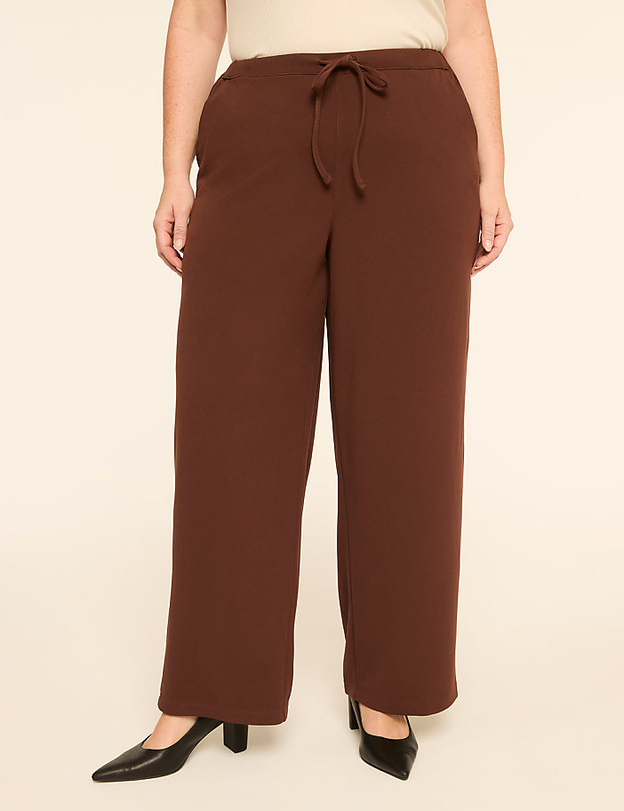 A person wears brown wide-leg trousers and black pointed heels against a plain, light-colored background.