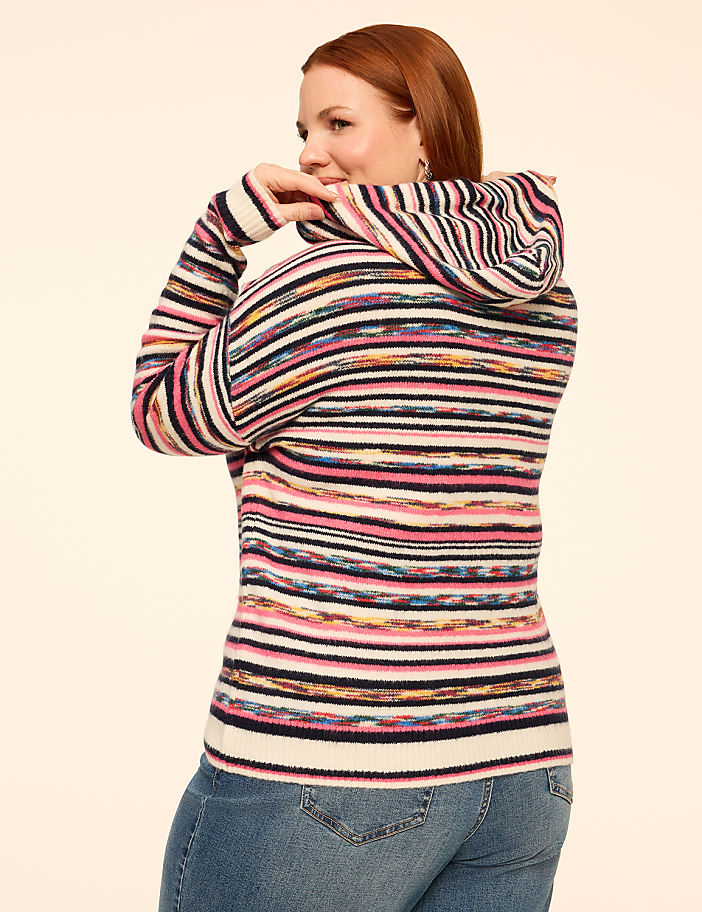 A woman with red hair wearing a colorful striped cowl-neck sweater and blue jeans stands against a plain, light-colored background.