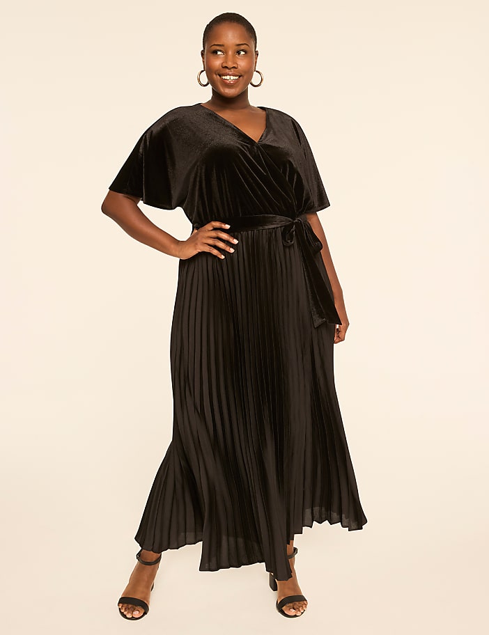 A smiling woman in a black velvet wrap dress with a pleated skirt stands against a plain, light-colored background.