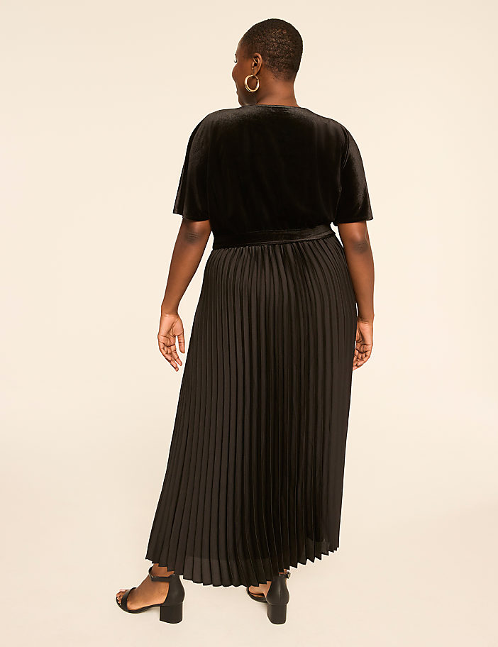 A woman in a black velvet top and pleated skirt stands against a plain, light-colored background.