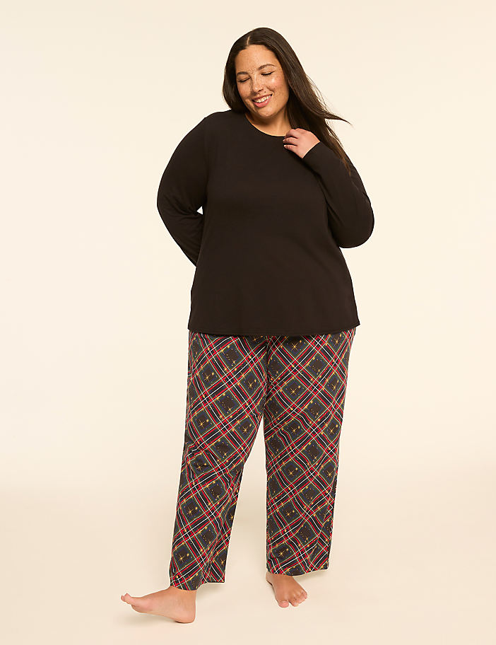 A smiling woman in a black top and plaid pajama pants stands on a plain, light-colored background.