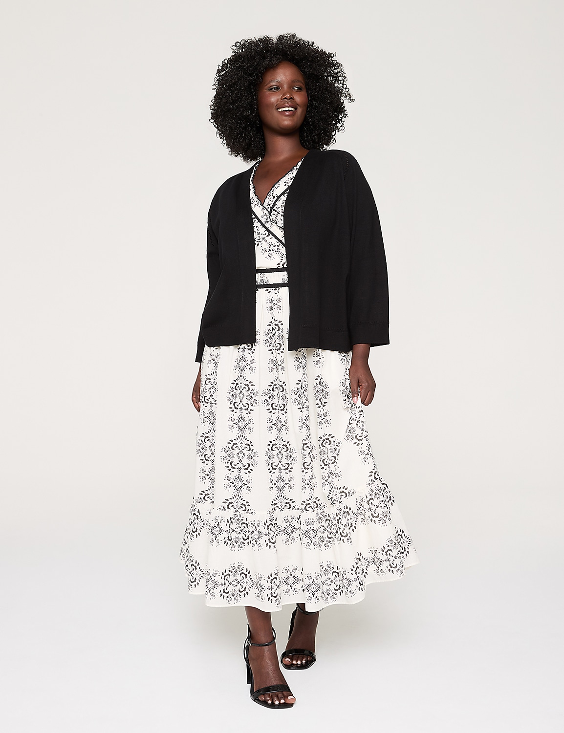 A smiling woman with curly black hair wearing a white floral dress and a black cardigan stands against a plain white background. Product Image 3