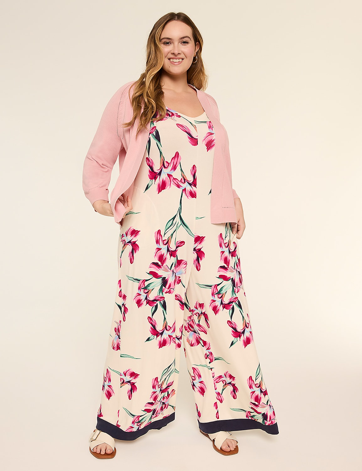 A young woman with long, wavy hair is wearing a pink jacket and a floral-patterned jumpsuit, standing against a plain beige background and smiling at the camera. Product Image 3