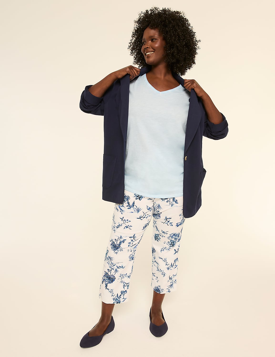 A smiling person with curly hair wearing a white shirt, navy blue jacket, and patterned pants stands against a plain background. Product Image 3
