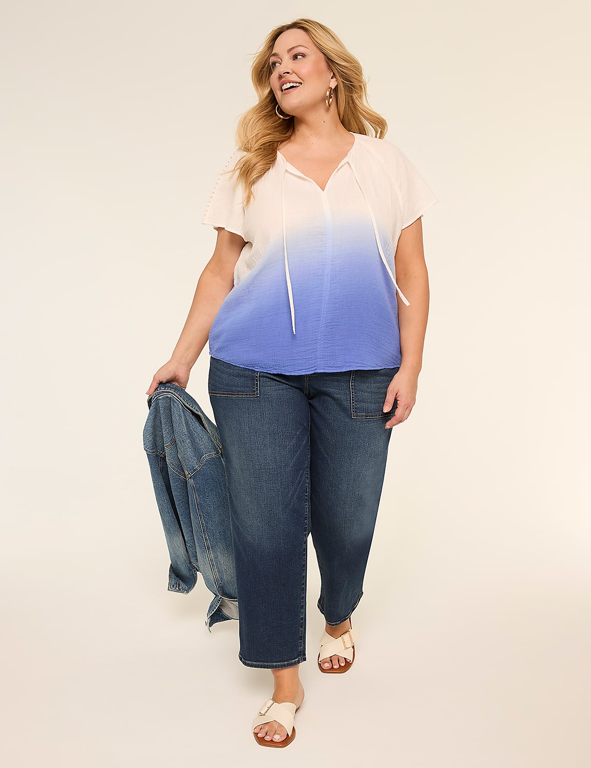 A smiling woman in a white and blue ombre top and dark jeans walks forward against a plain, light background, holding a denim jacket. Product Image 3