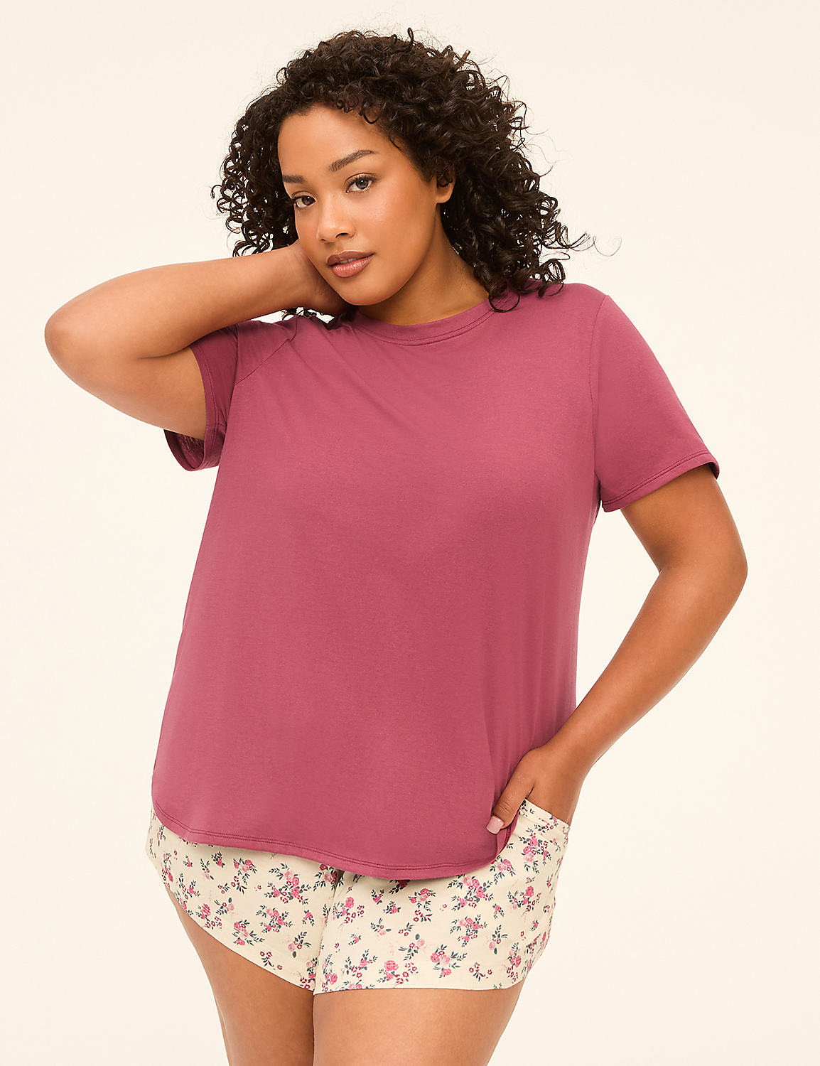 A woman with curly dark hair poses in a mauve t-shirt and floral shorts against a plain cream background. Product Image 1