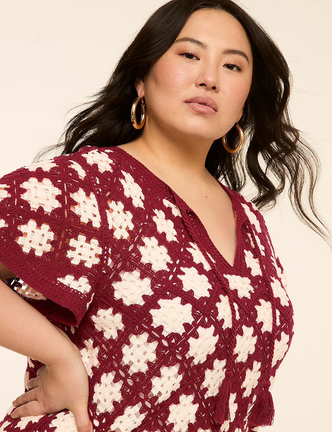 A woman with dark hair and gold hoop earrings poses in a maroon and cream crocheted top against a plain beige background. Product Image 4