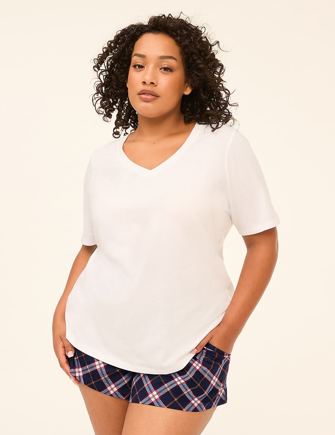 A woman with curly dark hair stands in the foreground wearing a white v-neck t-shirt and plaid shorts against a plain, light-colored background. Product Image 3