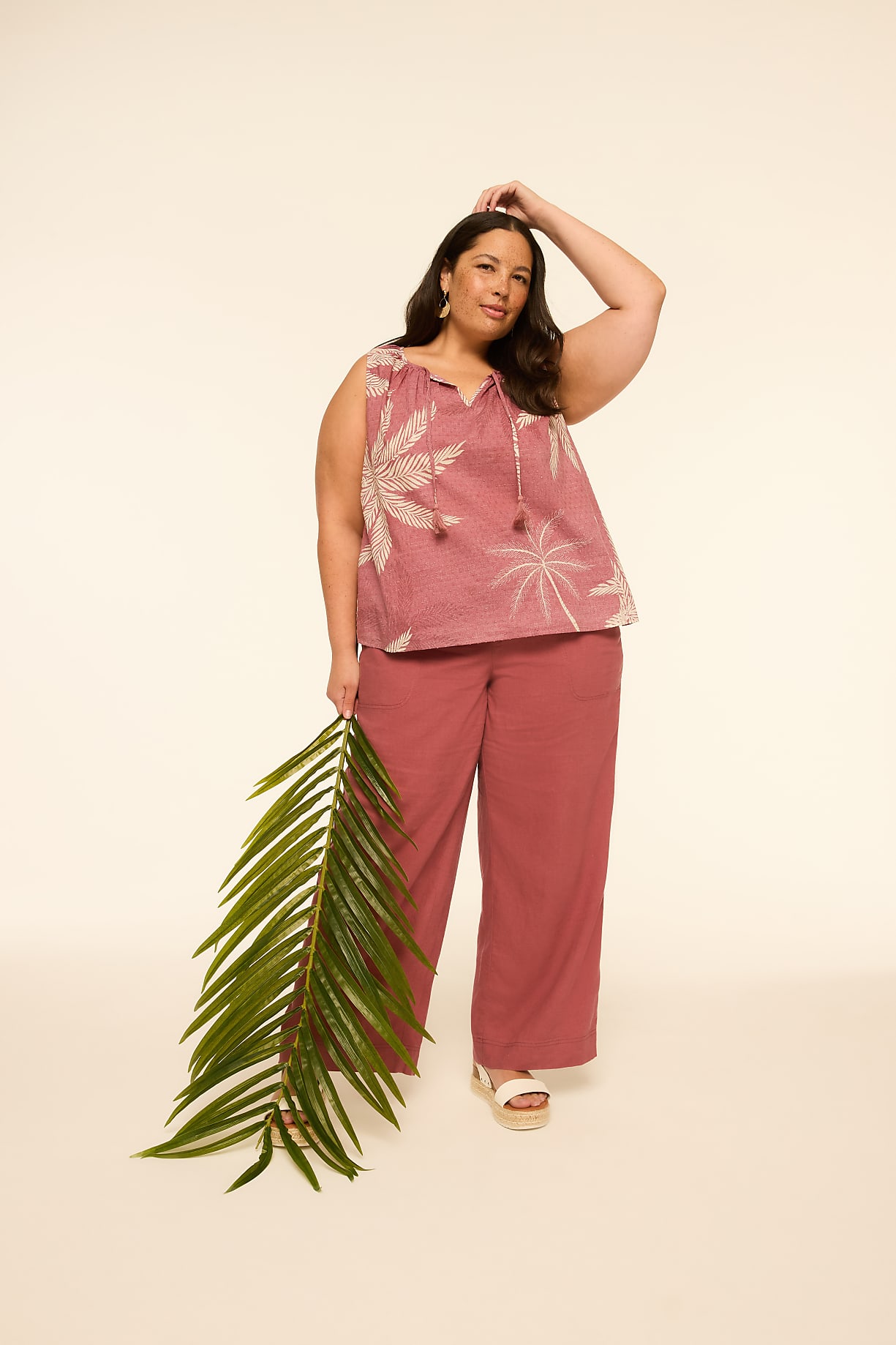 A woman in a pink patterned top and matching pants holds a large green palm frond in the foreground against a plain, light-colored background. Product Image 6