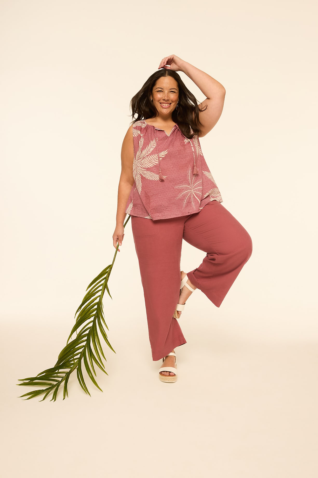 A smiling woman in a pink top and pants poses with a palm frond against a plain, light background. Product Image 10