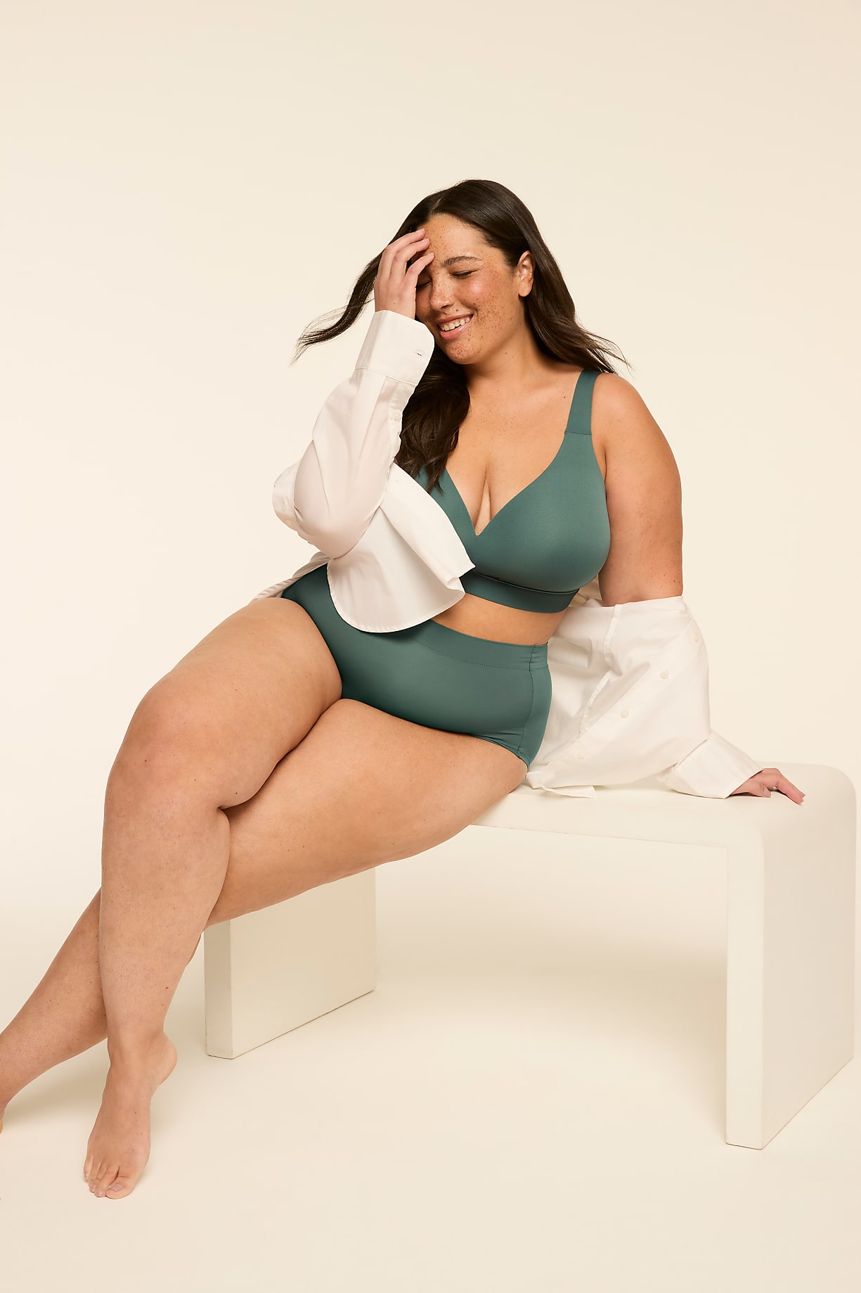 A smiling woman in teal lingerie and an open white shirt sits on a white bench against a plain cream background. Product Image 5