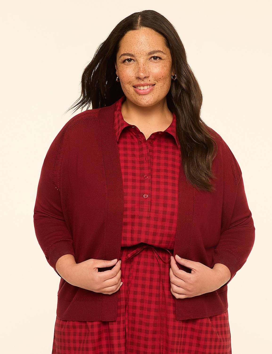 A smiling woman with dark hair and freckles, wearing a maroon cardigan over a red gingham dress, stands against a plain cream background. Product Image 1