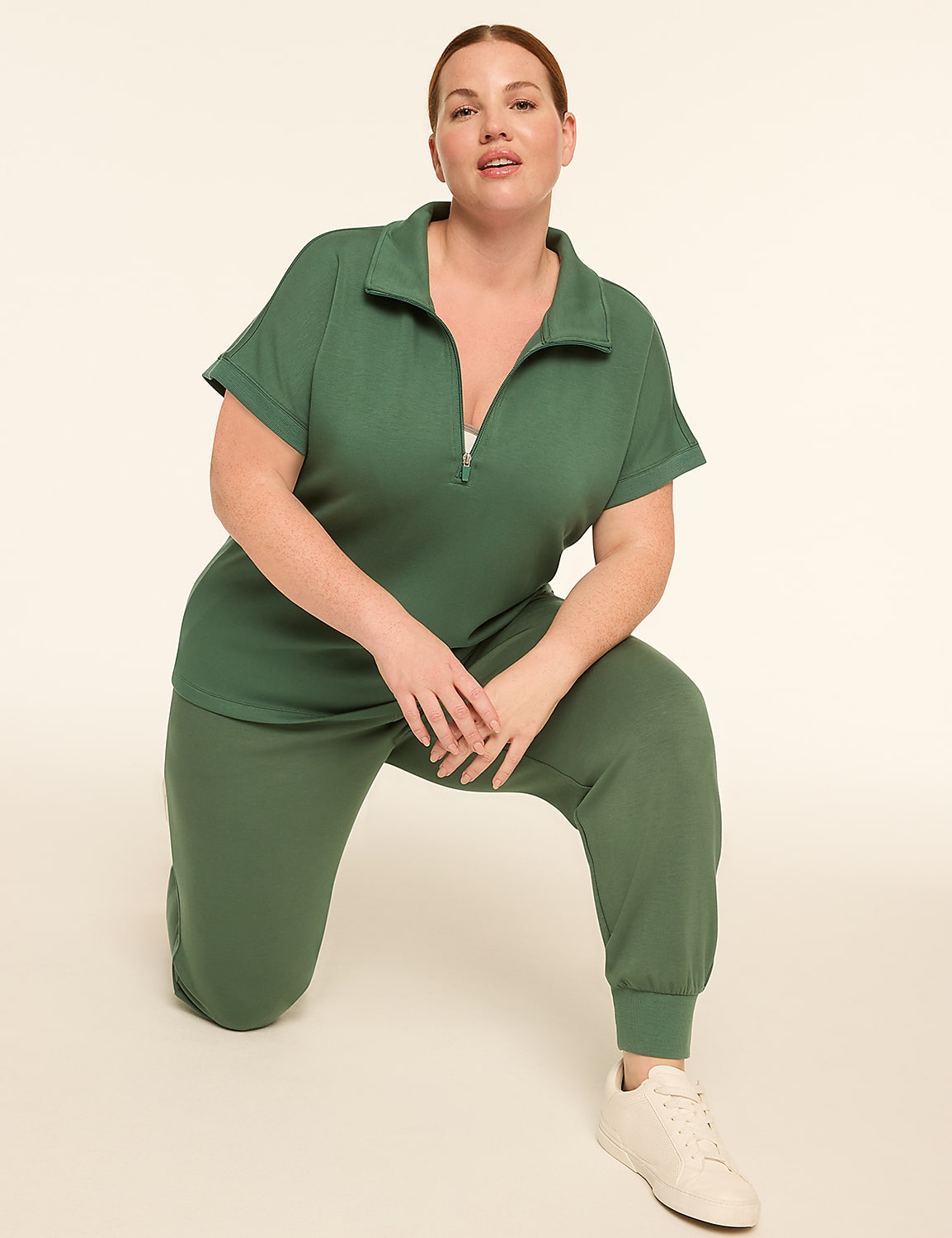 A woman in a green jumpsuit kneels on a light beige floor with a plain, light beige background. Product Image 3