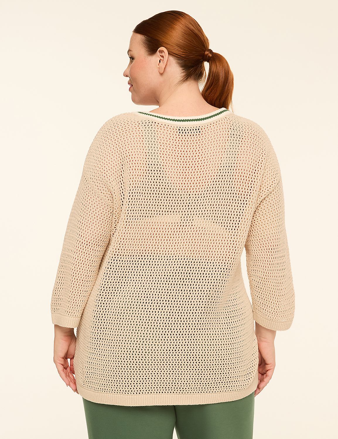 A woman with auburn hair pulled back in a ponytail stands facing away from the camera, wearing a loose-fitting, cream-colored, open-knit sweater with green trim at the neckline, and green pants, against a plain, light beige background. Product Image 2