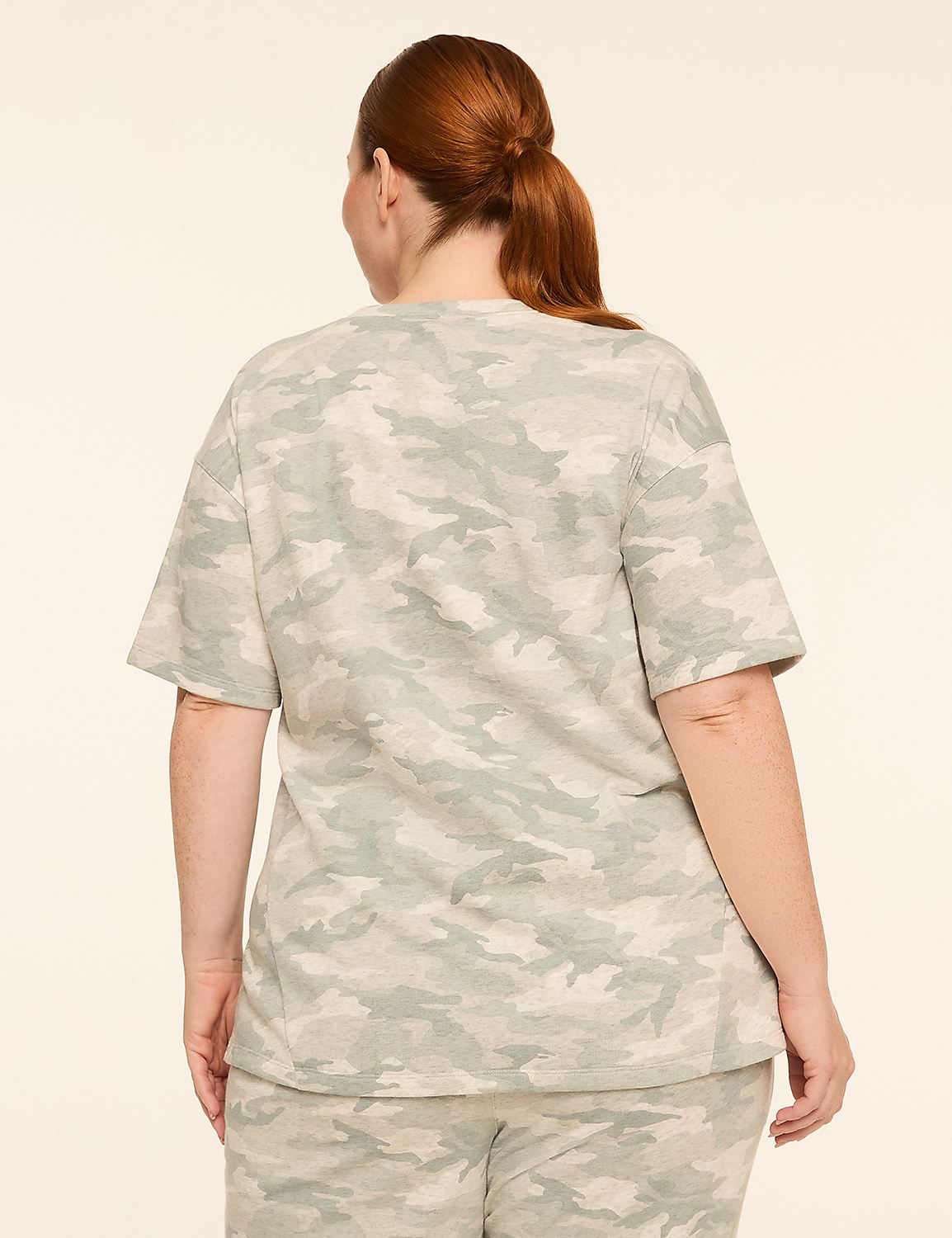 A woman with red hair pulled back in a ponytail stands facing away from the camera, wearing a camouflage t-shirt and pants against a plain, light-colored background. Product Image 2