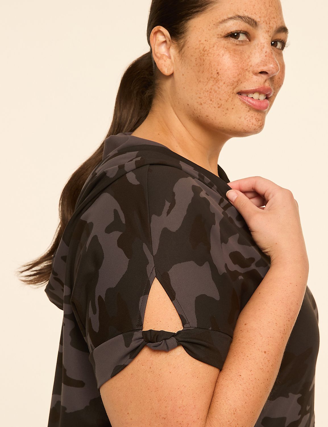 A woman with freckles smiles as she poses in a camouflage hoodie with knotted sleeves against a plain cream background. Product Image 3
