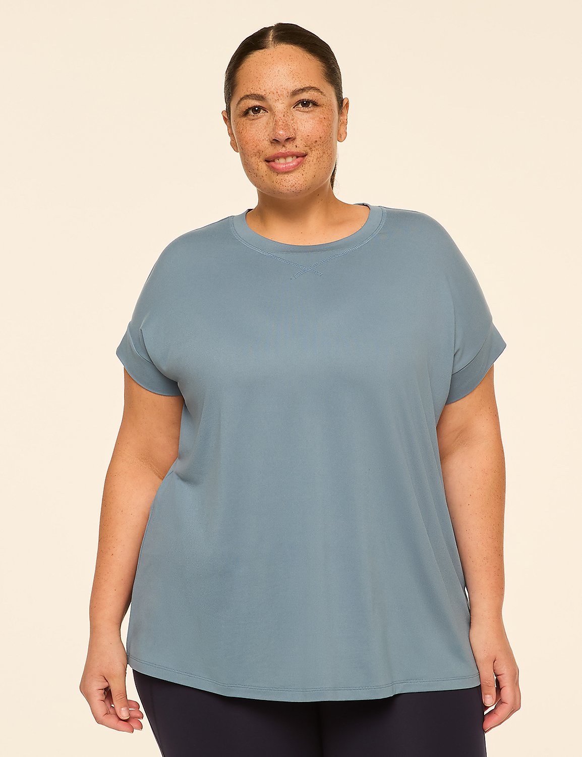 A woman with freckles smiles in a blue t-shirt and dark pants against a plain cream background. Product Image 1