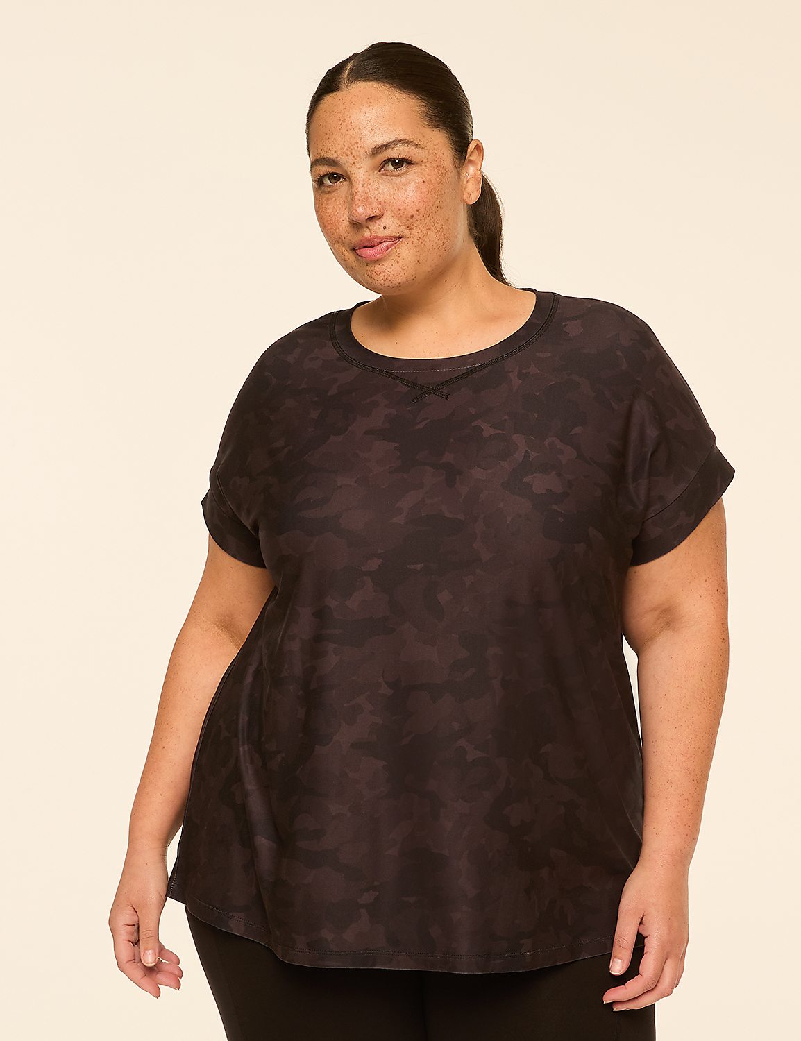 A woman with dark hair and freckles poses in a dark camouflage t-shirt against a plain, light-colored background. Product Image 1