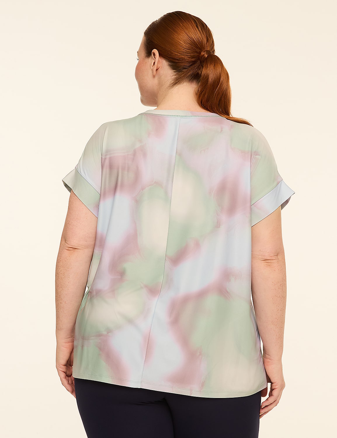 A woman with auburn hair pulled back in a ponytail stands facing away from the camera, wearing a light-colored, abstract-patterned blouse and dark pants against a plain, light beige background. Product Image 2