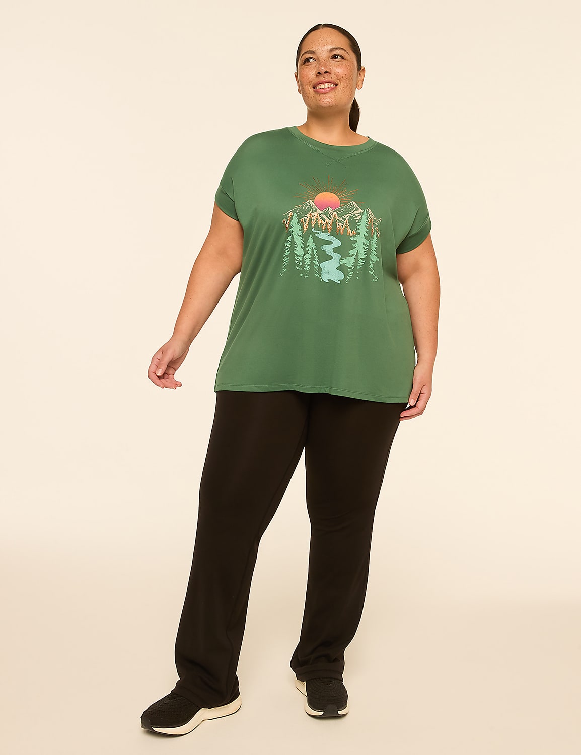 A woman in a green t-shirt with a mountain landscape graphic and black pants stands against a plain, light-colored background. Product Image 3