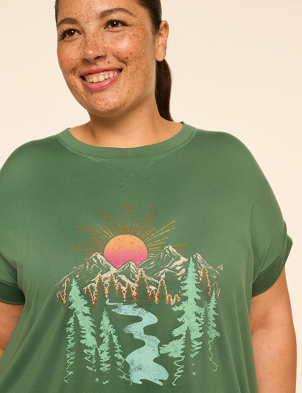 A smiling woman in a green t-shirt with a mountain and sun graphic stands against a plain background. Product Image 4