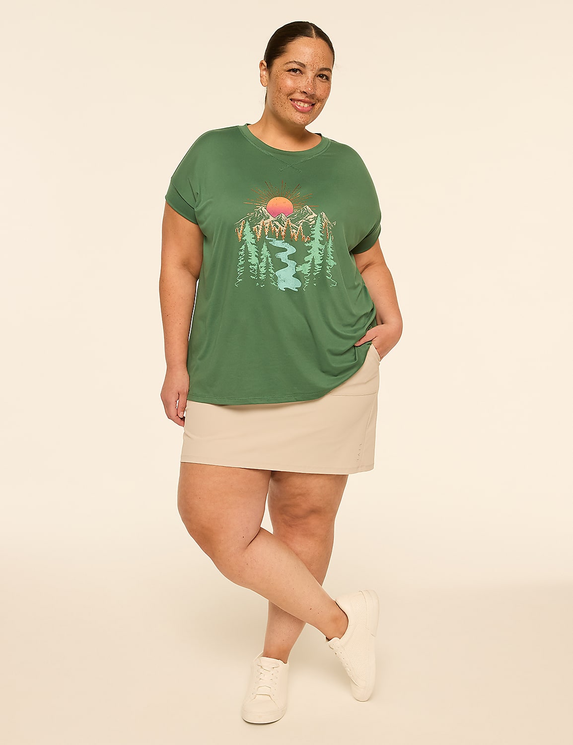 A woman in a green t-shirt with a mountain graphic and a tan skirt stands against a plain, light-colored background. Product Image 5