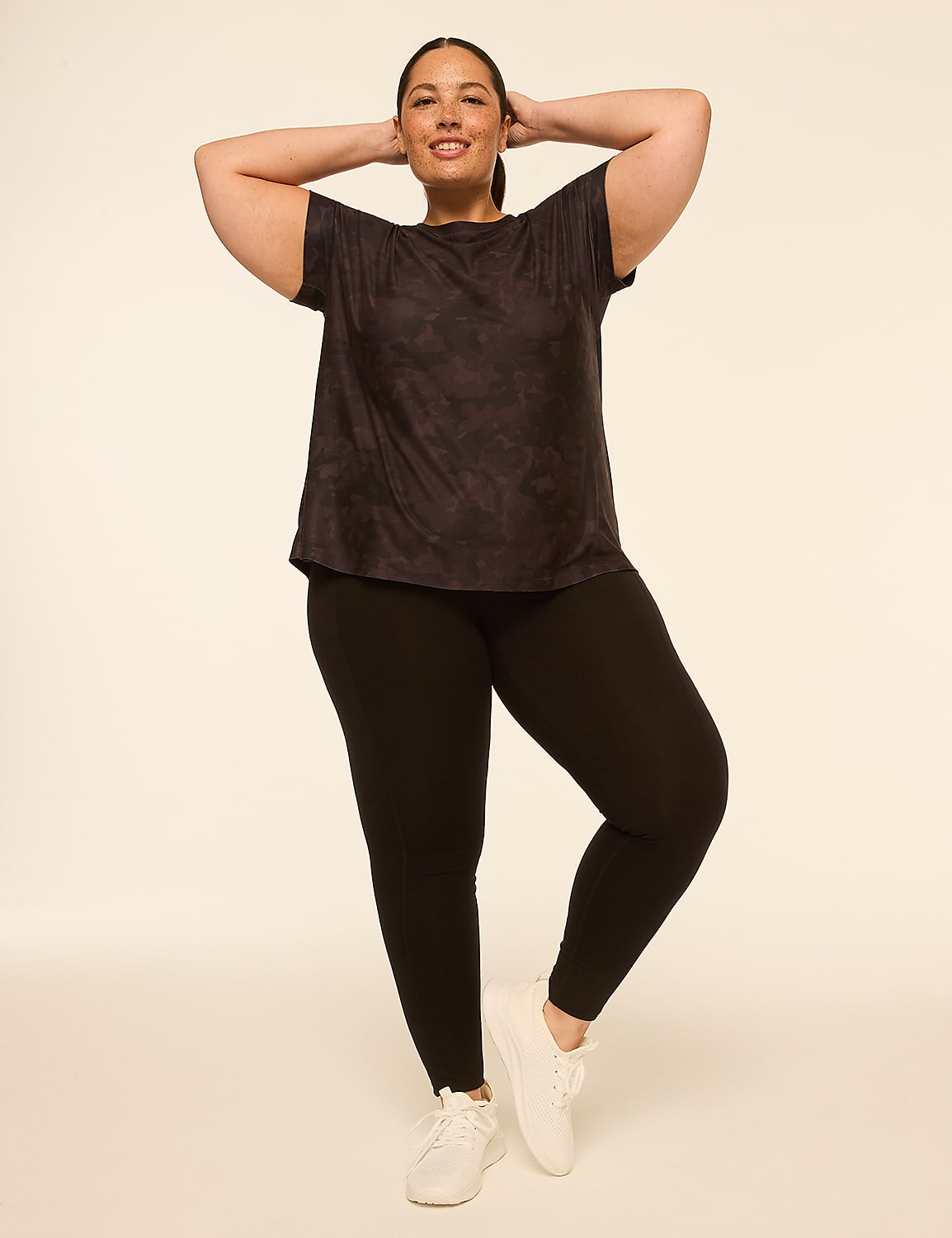 A woman in a dark tie-dye t-shirt and black leggings stands with her hands behind her head against a plain, light-colored background. Product Image 3