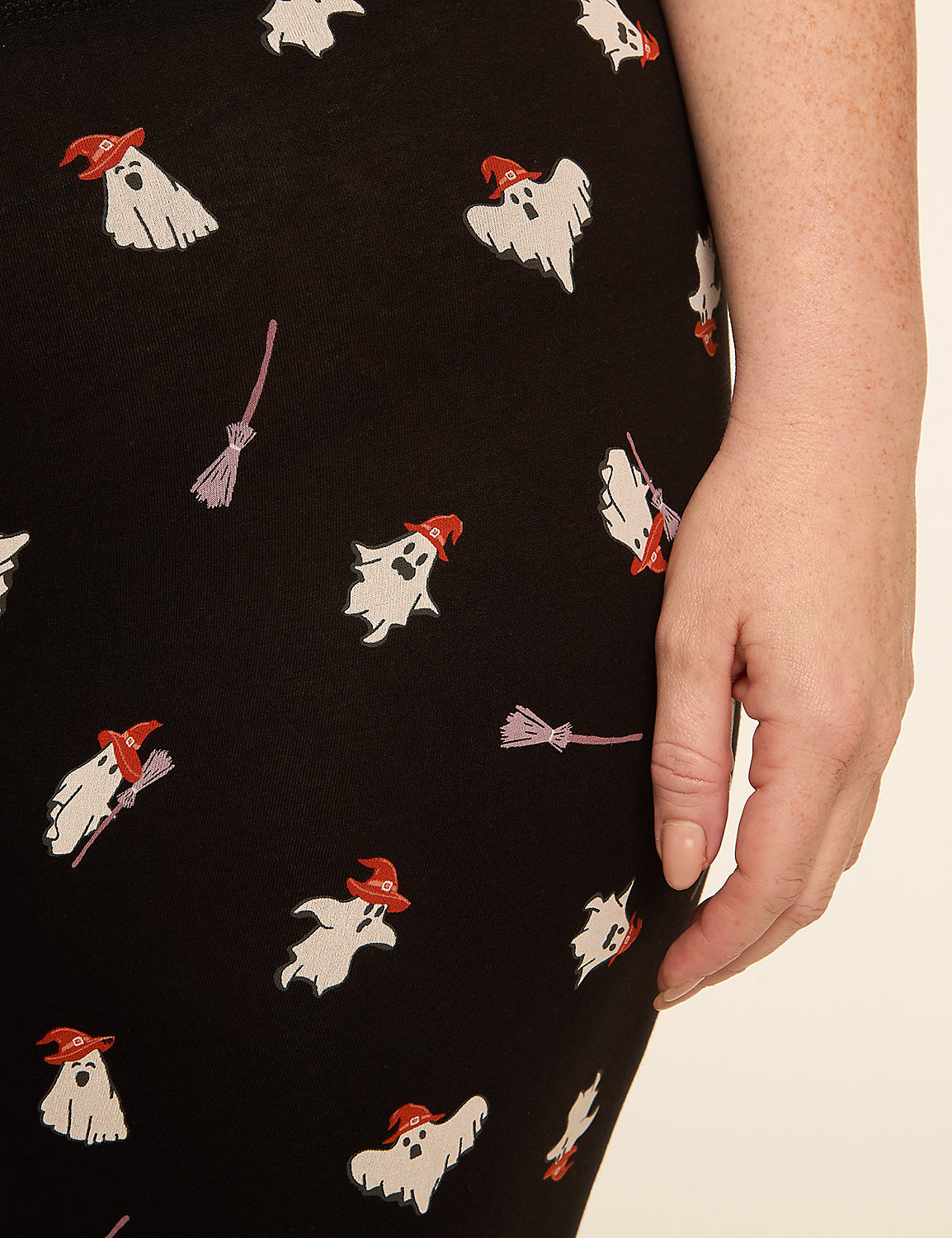 A woman's hand rests on black fabric patterned with white ghosts wearing orange hats and small purple brooms. Product Image 4