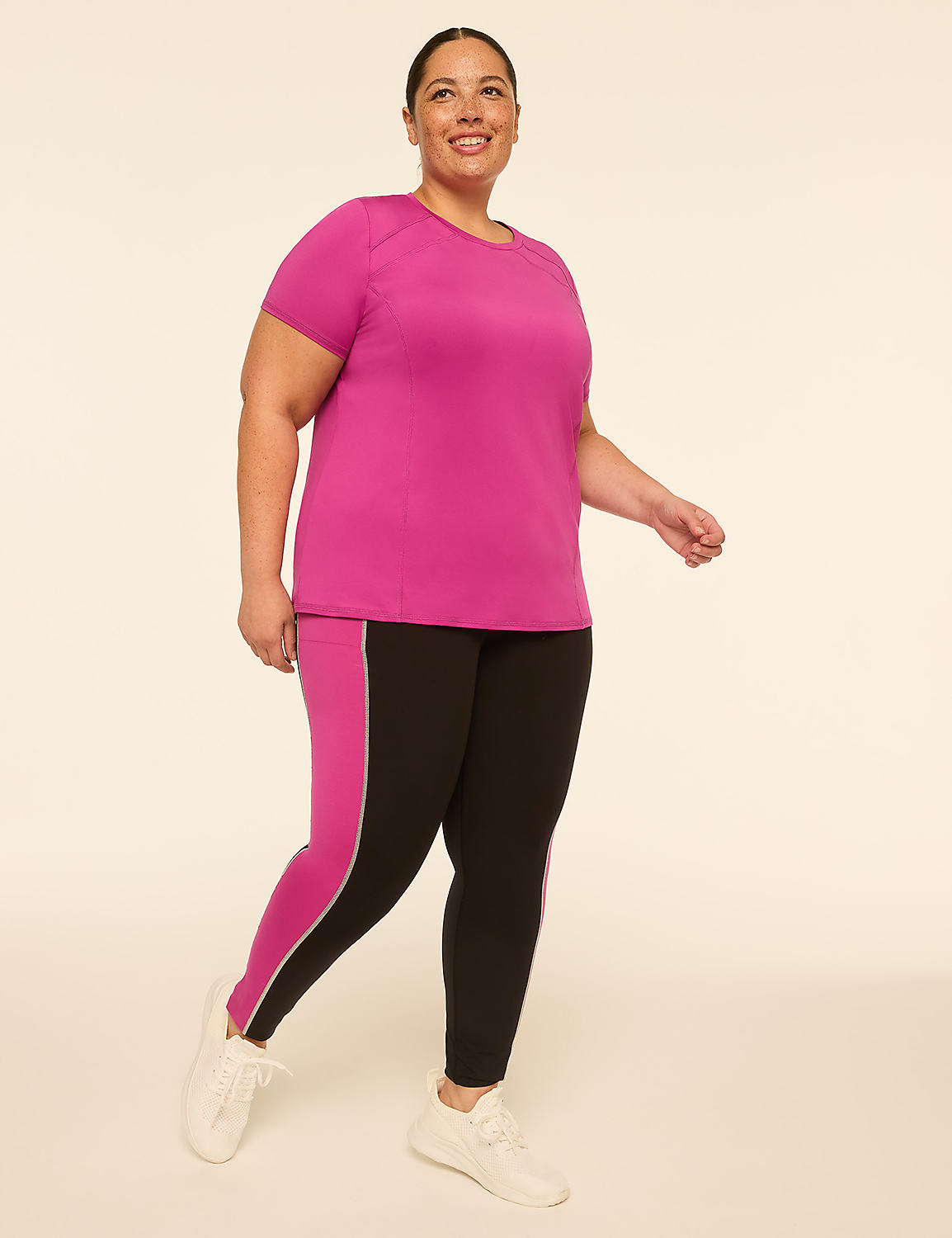 A smiling woman in a magenta t-shirt and black leggings with pink and white accents walks against a plain cream background. Product Image 3
