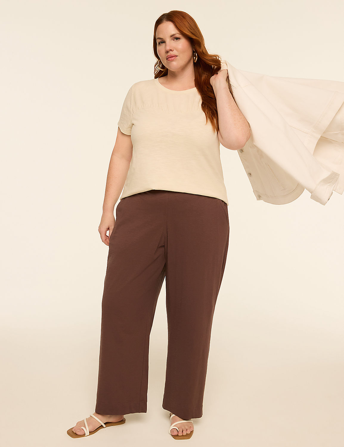 A woman in a cream top and brown pants stands against a plain, light background, holding a white jacket over her shoulder. Product Image 3