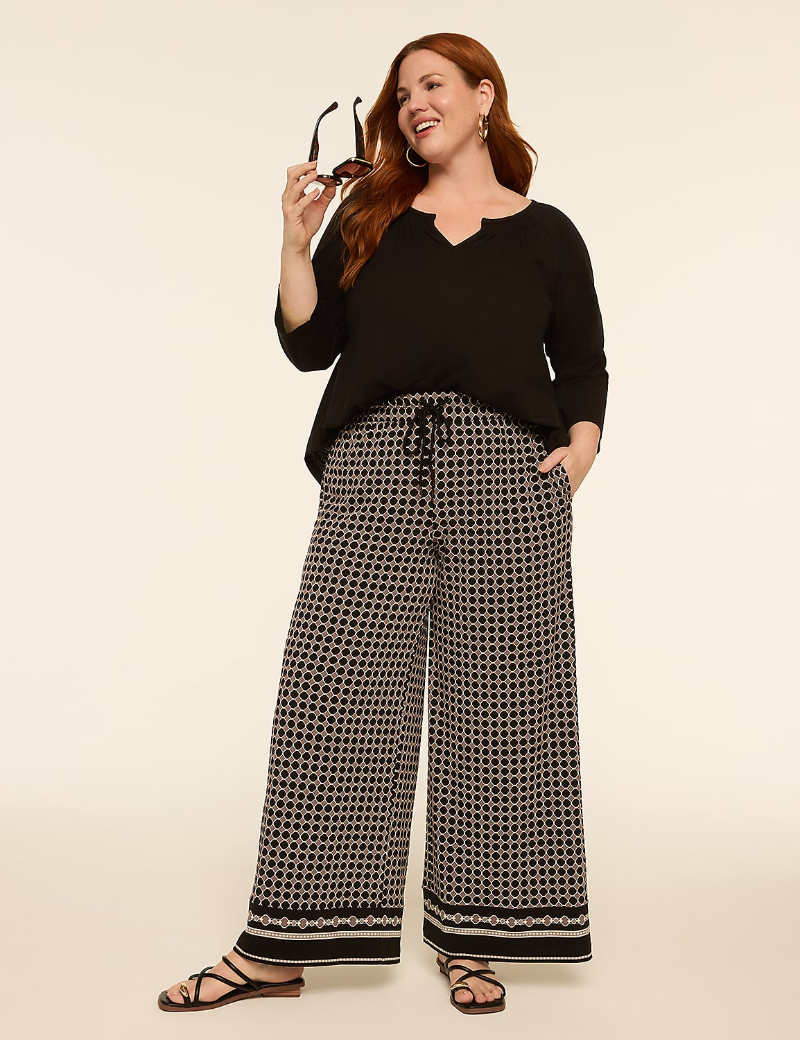 A woman with red hair stands in the foreground wearing a black top and patterned wide-leg pants, while the background is a plain, light-colored studio backdrop. Product Image 3