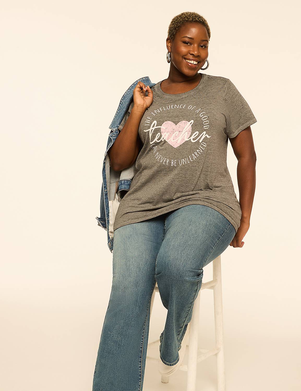 A smiling woman in a "teacher" t-shirt and jeans sits on a stool against a plain, light background. Product Image 3