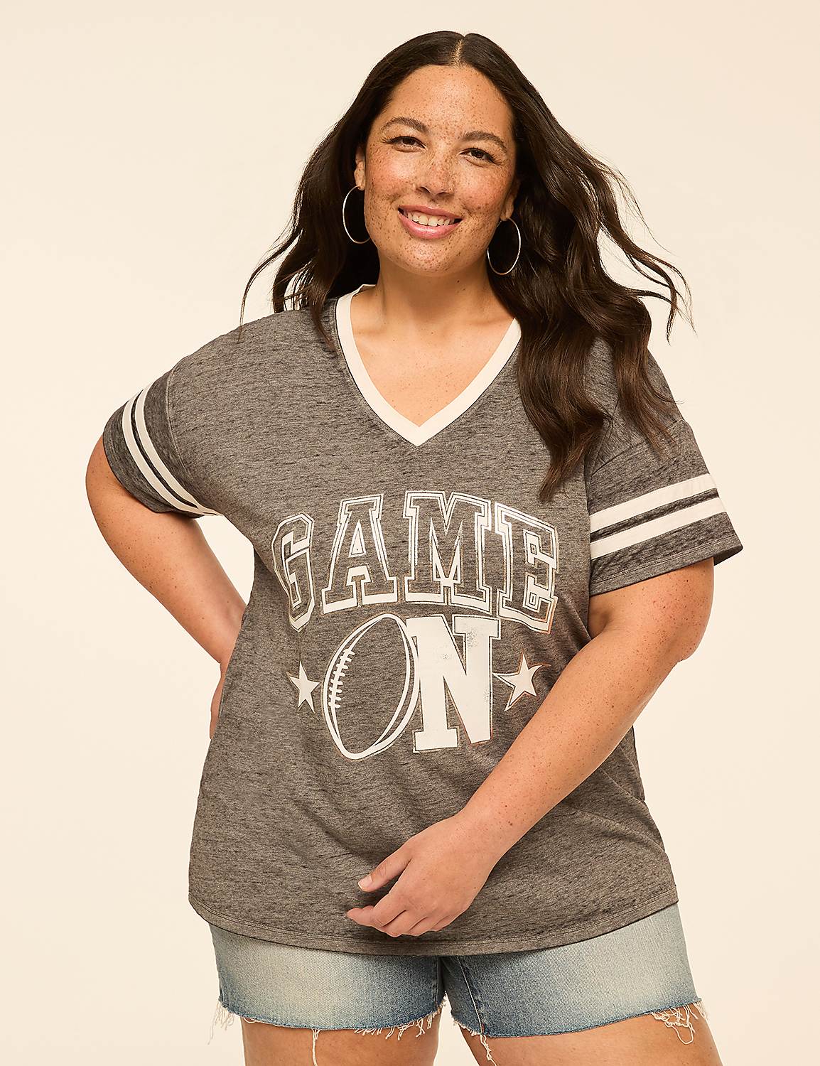 A smiling woman in a "GAME ON" t-shirt and denim shorts stands against a plain, light-colored background. Product Image 1