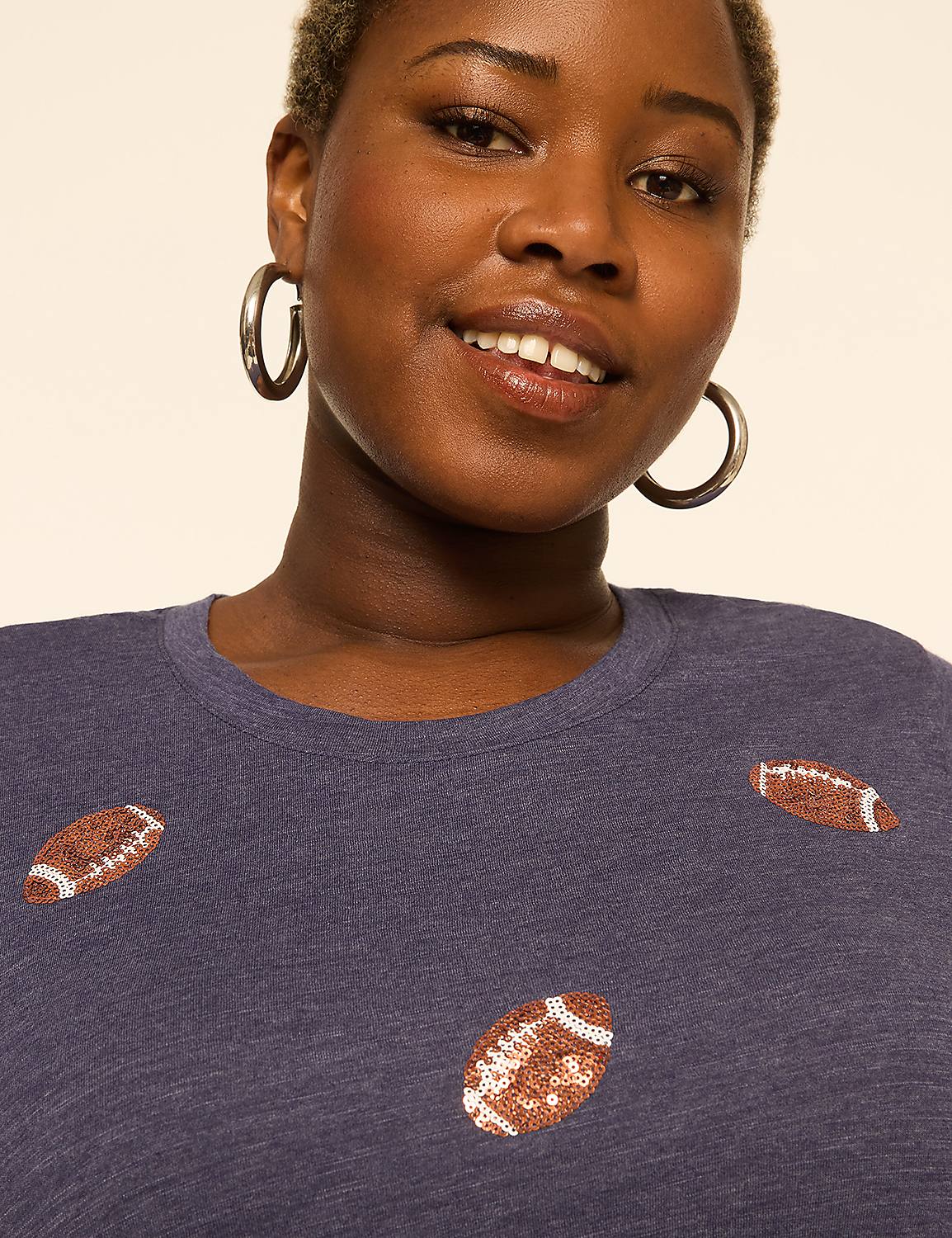 A smiling woman with short hair and hoop earrings wears a navy blue shirt adorned with footballs, set against a plain, light background. Product Image 4