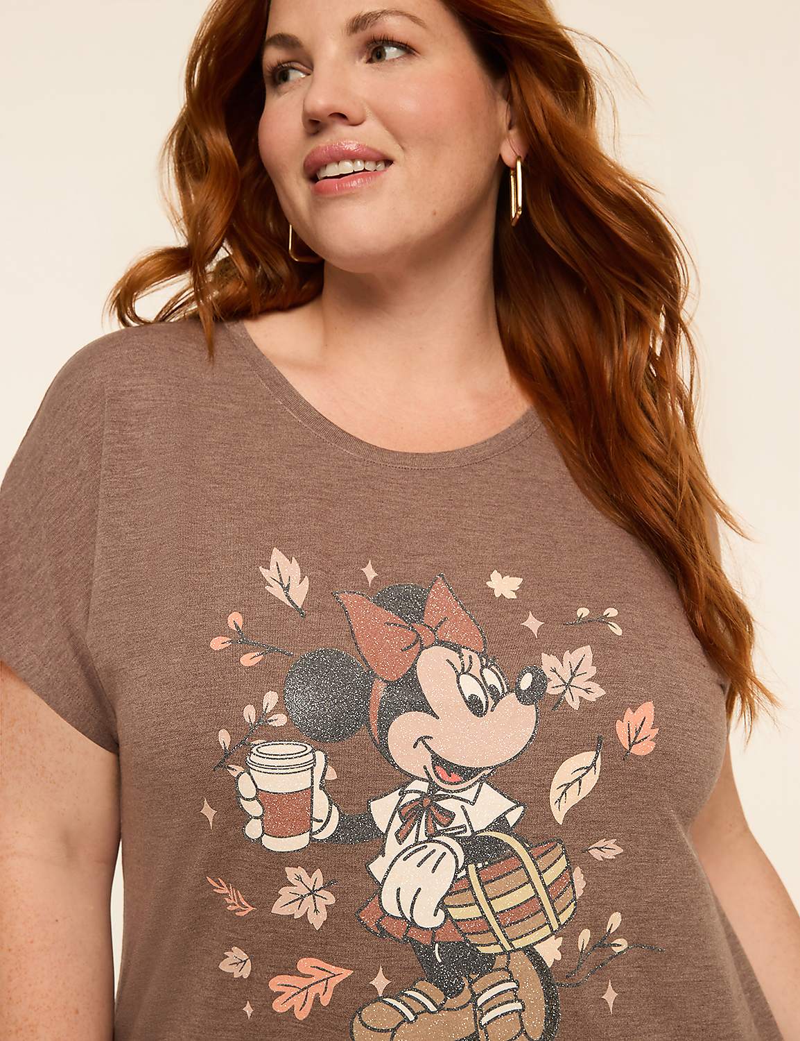 A woman with auburn hair smiles as she poses in front of a plain background, wearing a brown t-shirt with a graphic of Minnie Mouse holding a coffee cup and a basket surrounded by autumn leaves. Product Image 4