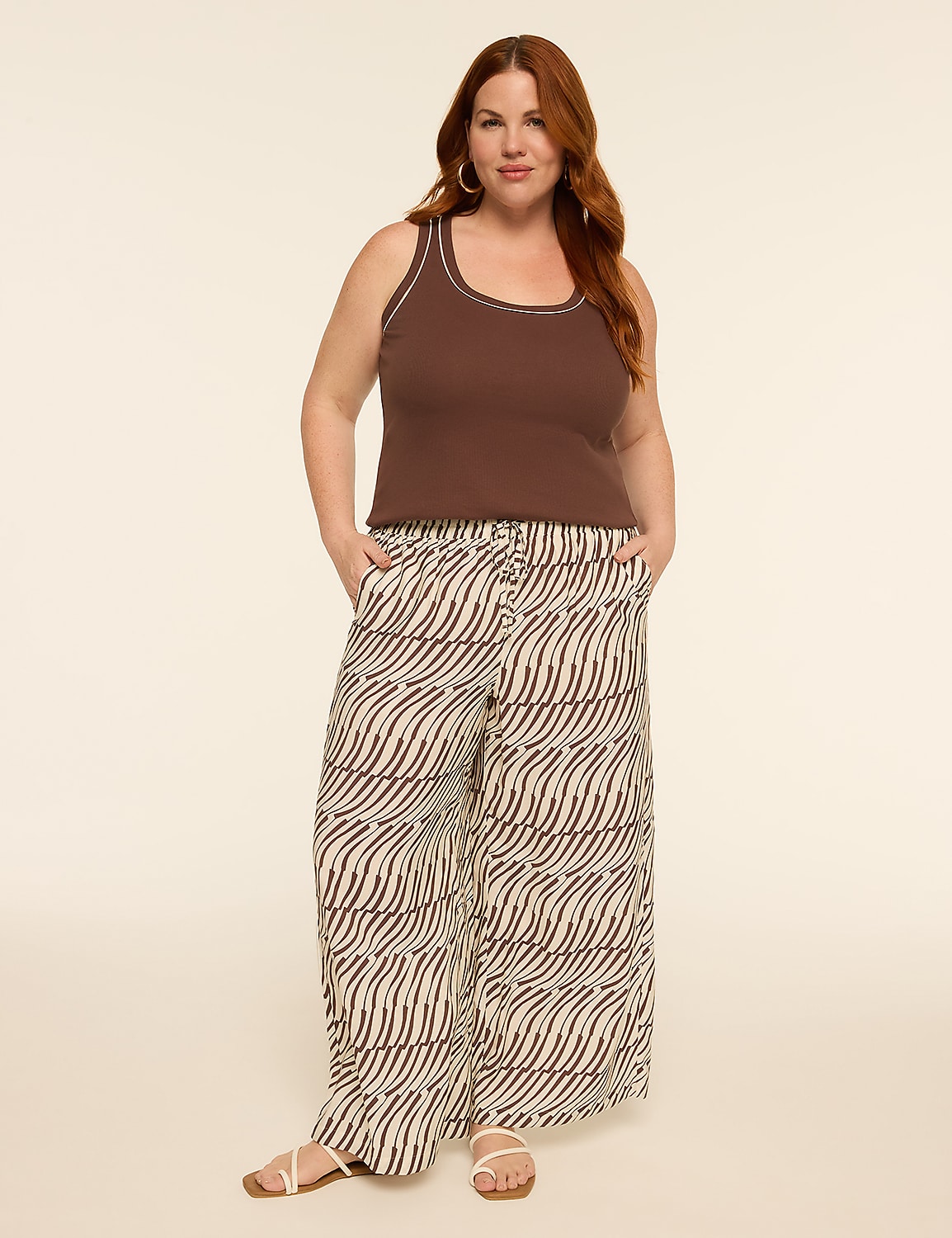 A woman with auburn hair stands in the foreground wearing a brown tank top and patterned wide-leg pants, against a plain, light-colored background. Product Image 3