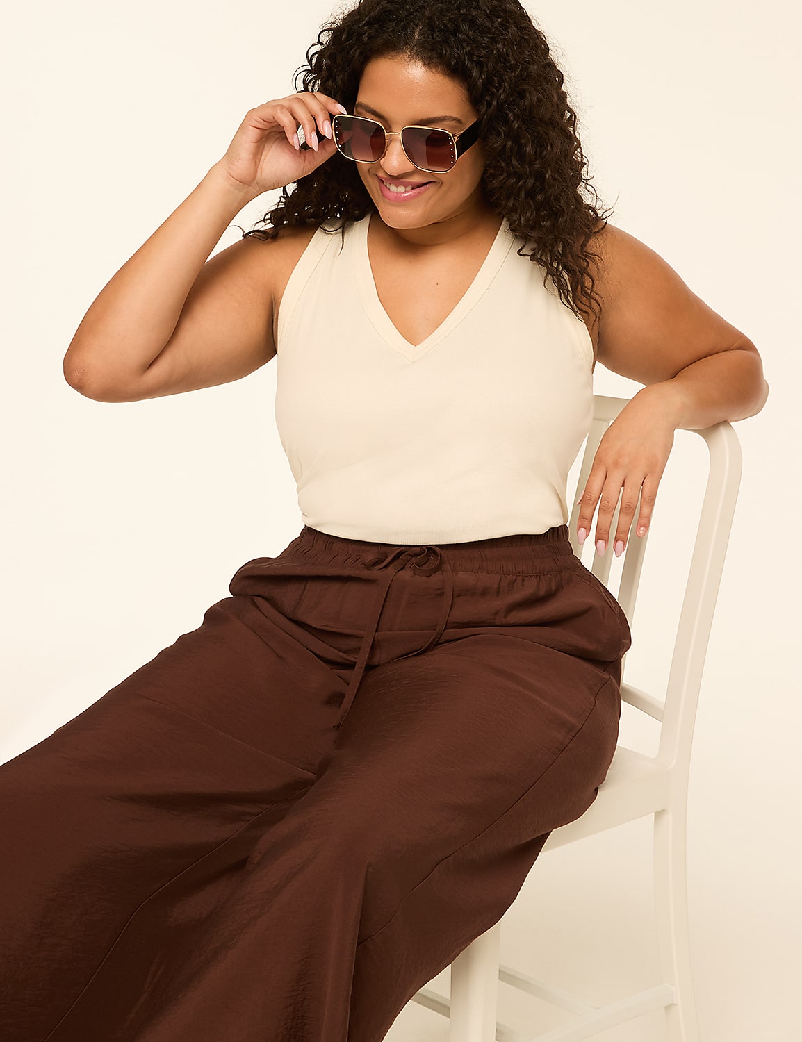 A woman in a cream v-neck top and brown wide-leg pants sits on a white chair, adjusting her sunglasses against a plain, light background. Product Image 3