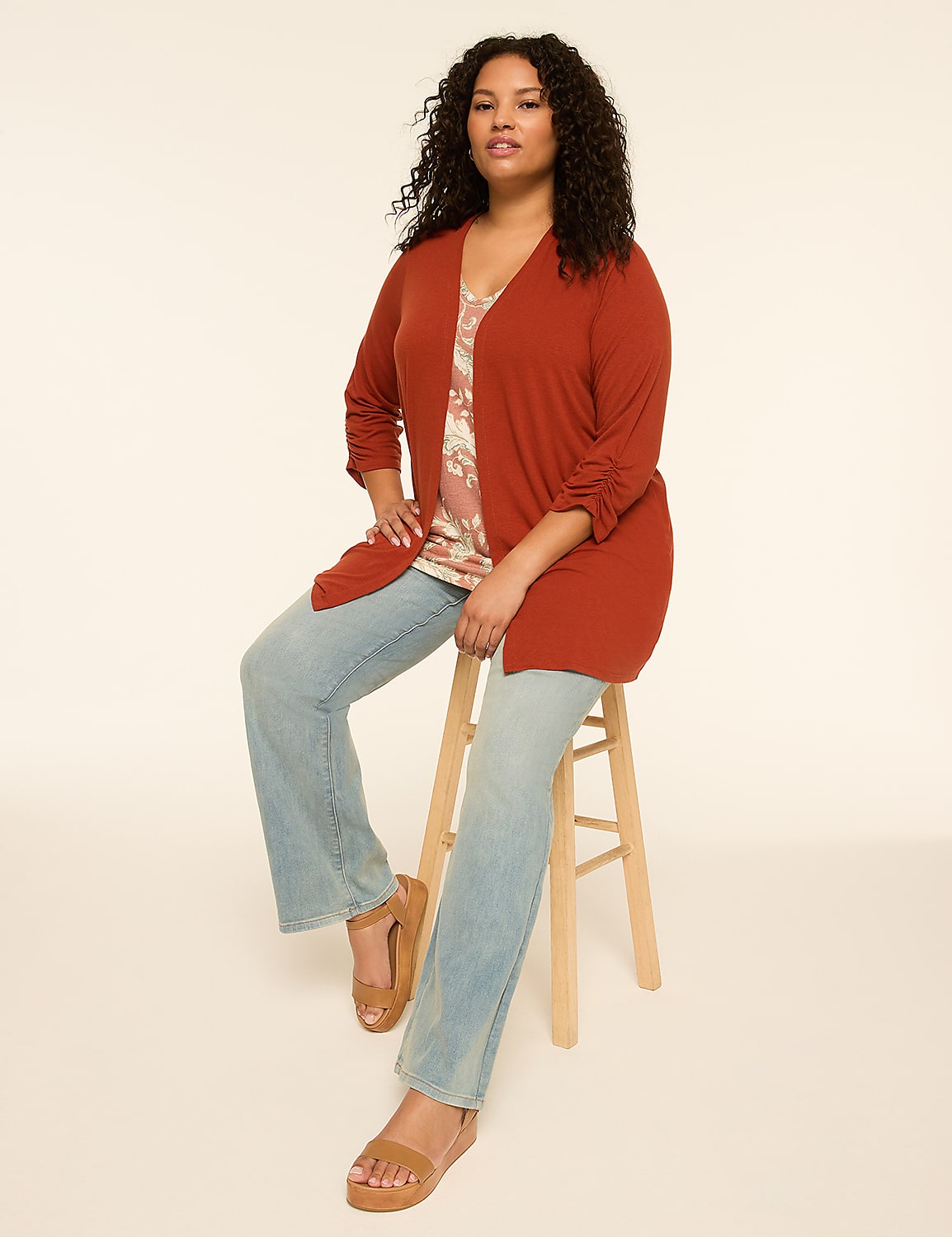 A woman with curly dark hair sits on a wooden stool wearing a rust-colored cardigan, a patterned top, and light-wash jeans against a plain, light background. Product Image 3