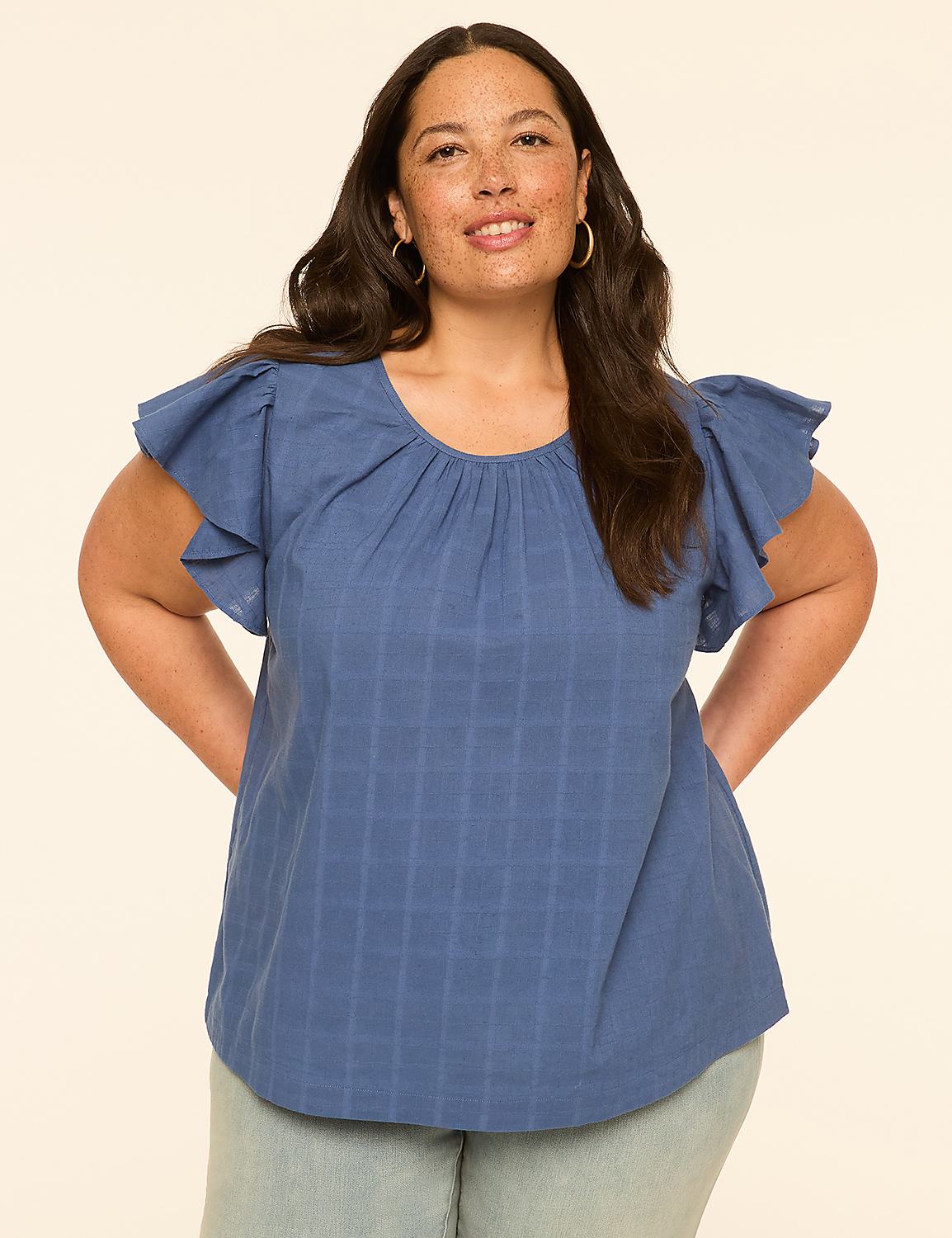 A smiling woman in a blue, ruffled-sleeve top stands against a plain, light-colored background. Product Image 1