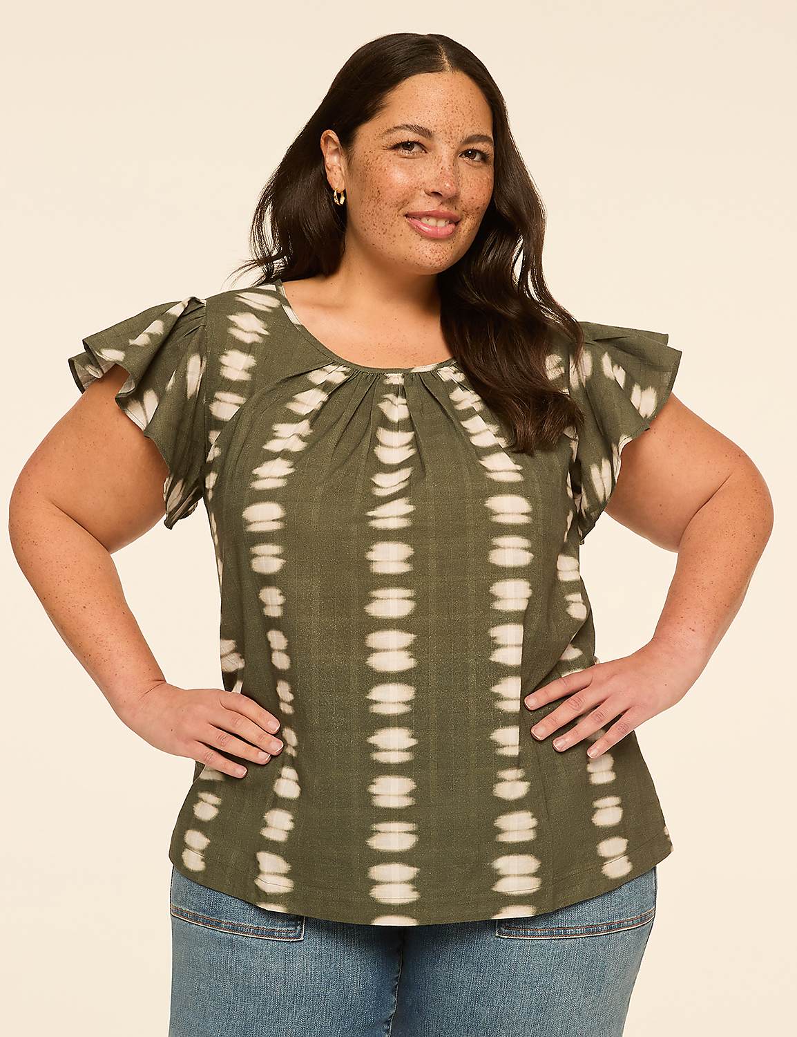 A woman with dark hair and a friendly smile stands with her hands on her hips, wearing a green tie-dye top and blue jeans against a plain, light-colored background. Product Image 1