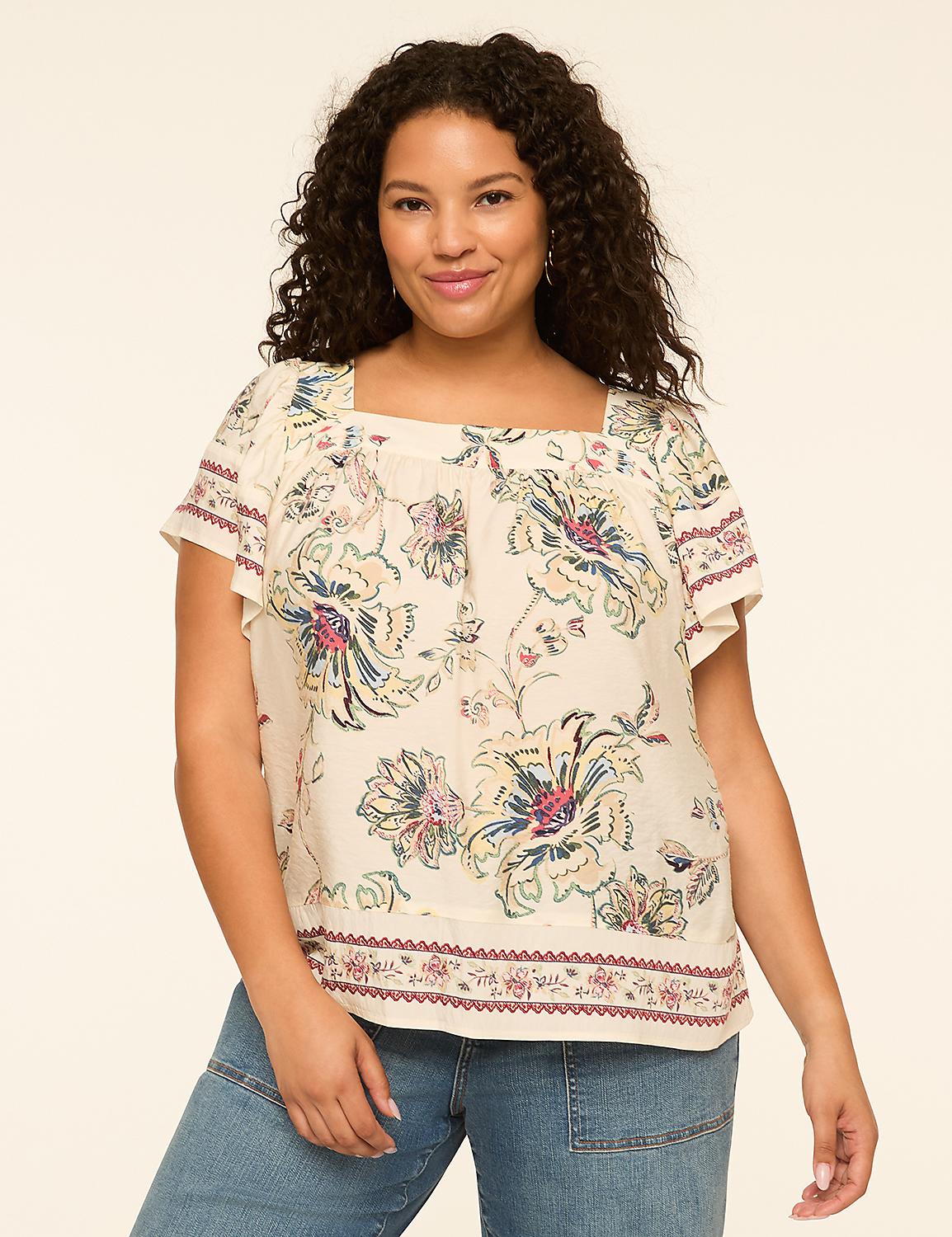 A smiling woman with curly dark hair wears a cream-colored floral print blouse with a square neckline and blue jeans, standing against a plain beige background. Product Image 1