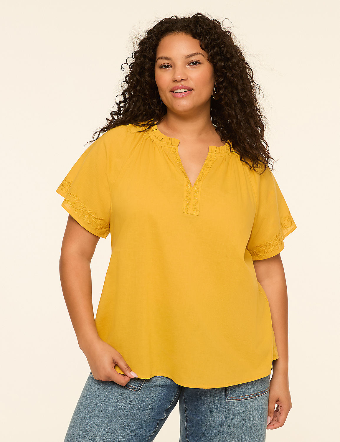 A woman with curly dark hair and a warm smile poses in a mustard yellow top and blue jeans against a plain, light beige background. Product Image 1