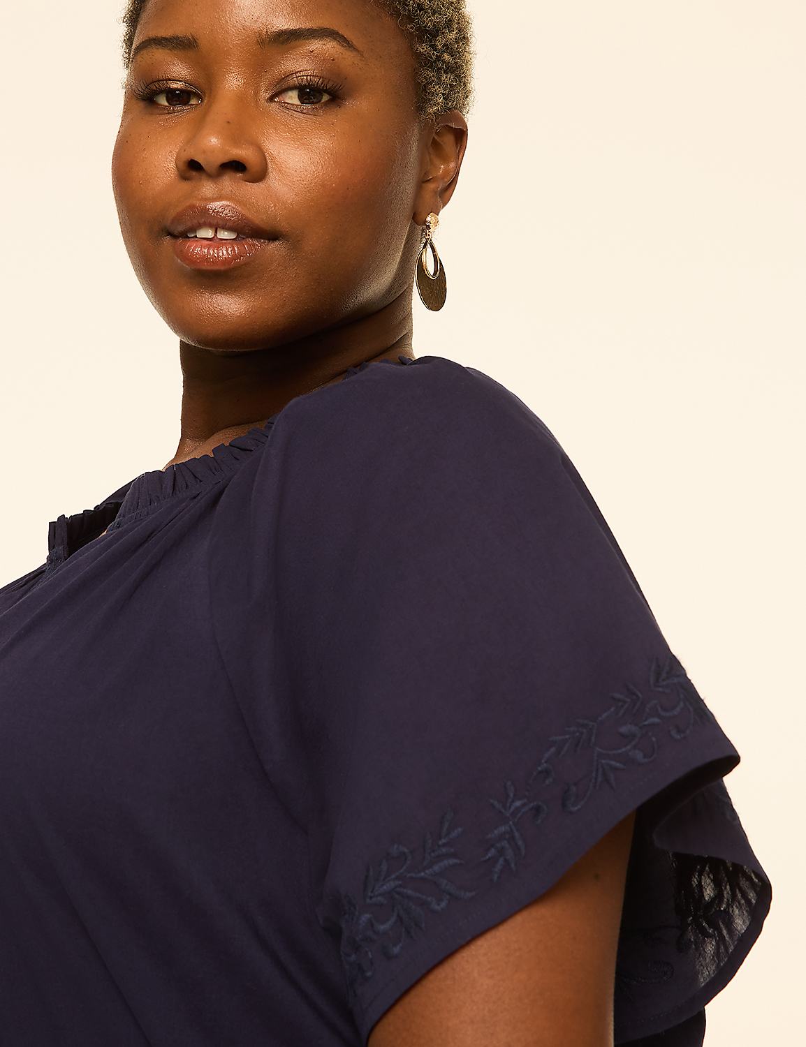 A woman with short, light-colored hair and a gold earring is shown in the foreground, wearing a navy blue top with embroidered sleeves, against a plain, light-colored background. Product Image 4