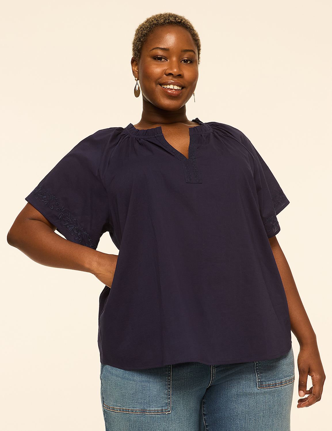 A smiling woman in a navy blue top and jeans stands against a plain, light-colored background. Product Image 1